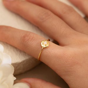 Puede incluir: Un delicado anillo de oro con una pequeña gema redonda y clara engastada en un diseño de estrella. El anillo está en un dedo, mostrando su estética simple pero elegante. La banda del anillo es delgada y pulida.