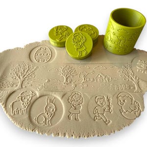 May include: A set of Easter-themed play dough stamps and a green rolling pin. The stamps include images of an Easter egg, a girl, a boy, a lamb, a bunny, and a chick. The play dough has a landscape design.