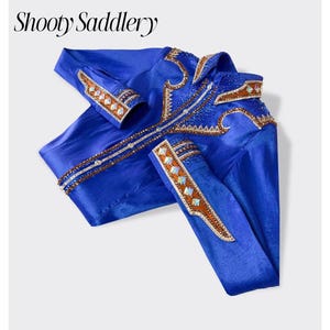 Peut inclure: Une chemise à manches longues bleu roi vif avec le texte Shooty Saddlery en haut. La chemise est ornée de garnitures décoratives marron et dorées complexes, d'accents en cristal clair et de motifs géométriques sur le col et les poignets.
