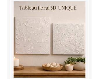 May include: Two white, textured floral 3D wall art pieces. The artwork features raised floral designs on a square canvas. The text "Tableau floral 3D UNIQUE" is at the top. A candle, a basket of decorative balls, and a potted plant are on a wooden surface.