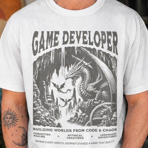Könnte beinhalten: Ein weißes T-Shirt mit einer dunkelgrauen Grafik eines Drachen in einer Höhle und dem Text GAME DEVELOPER, BUILDING WORLDS FROM CODE & CHAOS, FORGOTTEN REALMS, MYTHICAL CREATURES, LEGENDARY ADVENTURES und BEHIND EVERY HERO'S JOURNEY STANDS A MIND THAT BUILT IT.