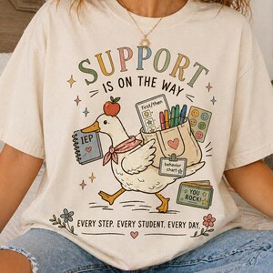 May include: Cream-colored t-shirt with the text "SUPPORT IS ON THE WAY" and a cartoon duck carrying school supplies. The duck has an apple on its head and a scarf. The shirt also has the text "EVERY STEP. EVERY STUDENT. EVERY DAY."