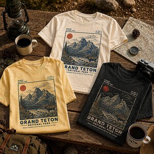 May include: Three short-sleeve t-shirts in cream, yellow, and black, each featuring a graphic print of Grand Teton National Park. The design includes a mountain range, a lake, a moose, a red sun, and the text "Grand Teton National Park."