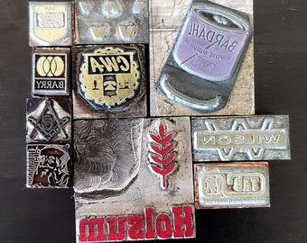 May include: A collection of vintage metal printing blocks with various designs and text, including "BARRY", "CWA", "WILSON", "HOLSUM", and "ADDITIVE MOTOR OIL BARDAHL".