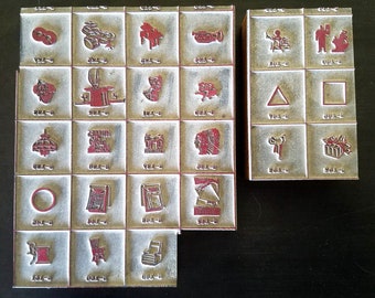 May include: A set of 25 vintage wooden stamps with red ink pads. The stamps feature various designs, including a musical instrument, a triangle, a square, a house, a lamp, a book, a hat, a bird, a gift, and a circle. The stamps are arranged in a grid pattern on a wooden surface.