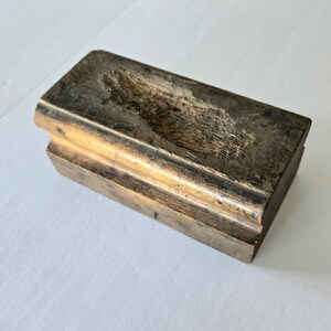 May include: A rectangular, antique wooden block with a carved groove and a curved edge. The wood has a weathered, aged appearance with shades of brown and gold, suggesting it's an old tool or decorative item.