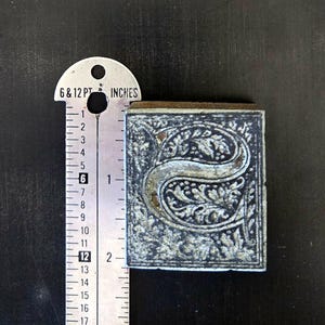 May include: A vintage letterpress printing block featuring the letter "S" in a decorative, ornate font. The block is made of metal and has a dark gray finish. The letter "S" is surrounded by a floral design.