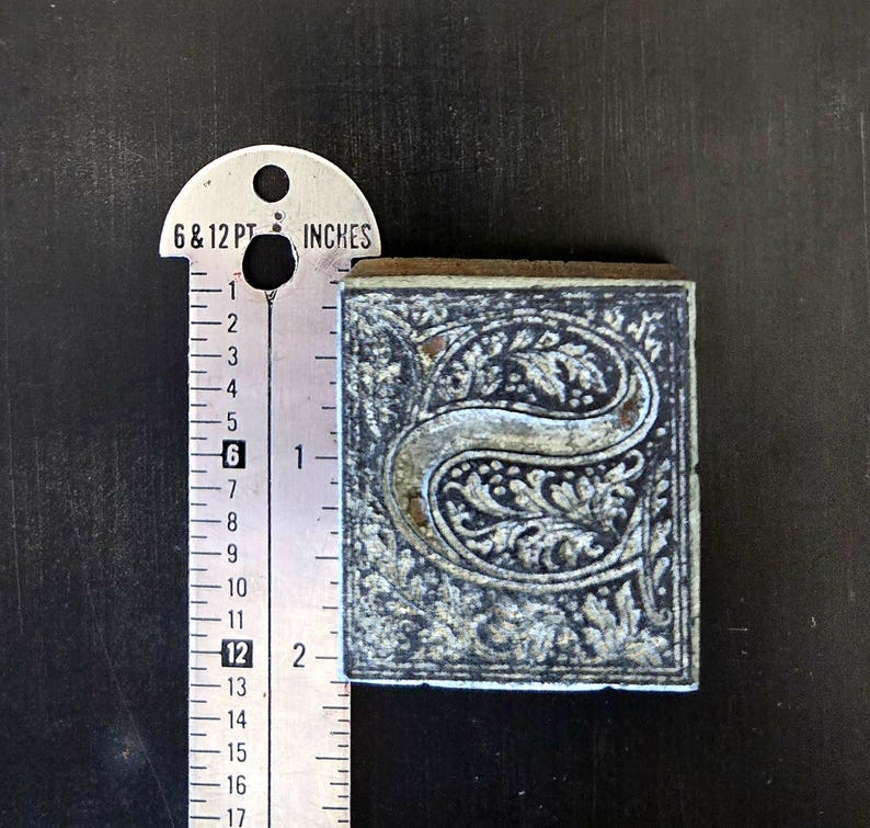 May include: A vintage letterpress printing block featuring the letter "S" in a decorative, ornate font. The block is made of metal and has a dark gray finish. The letter "S" is surrounded by a floral design.