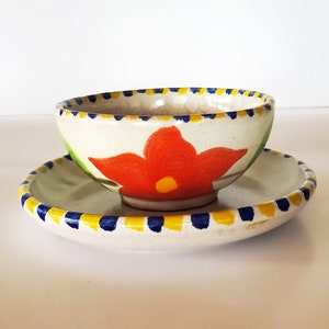 May include: A white ceramic bowl with a red flower design and blue and yellow trim. The bowl is sitting on a matching saucer.