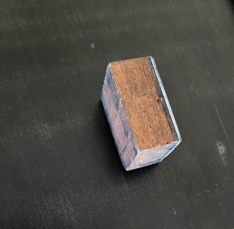 May include: A wooden block with a light brown color and a dark brown wood grain. The block has a thin silver metal trim around the edges.