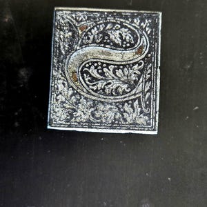 May include: A close-up of a silver metal stamp with an ornate letter "S" design. The stamp is on a black background with the text "BLACK BEAUTY TRADE MARK REG. U.S. 57106".