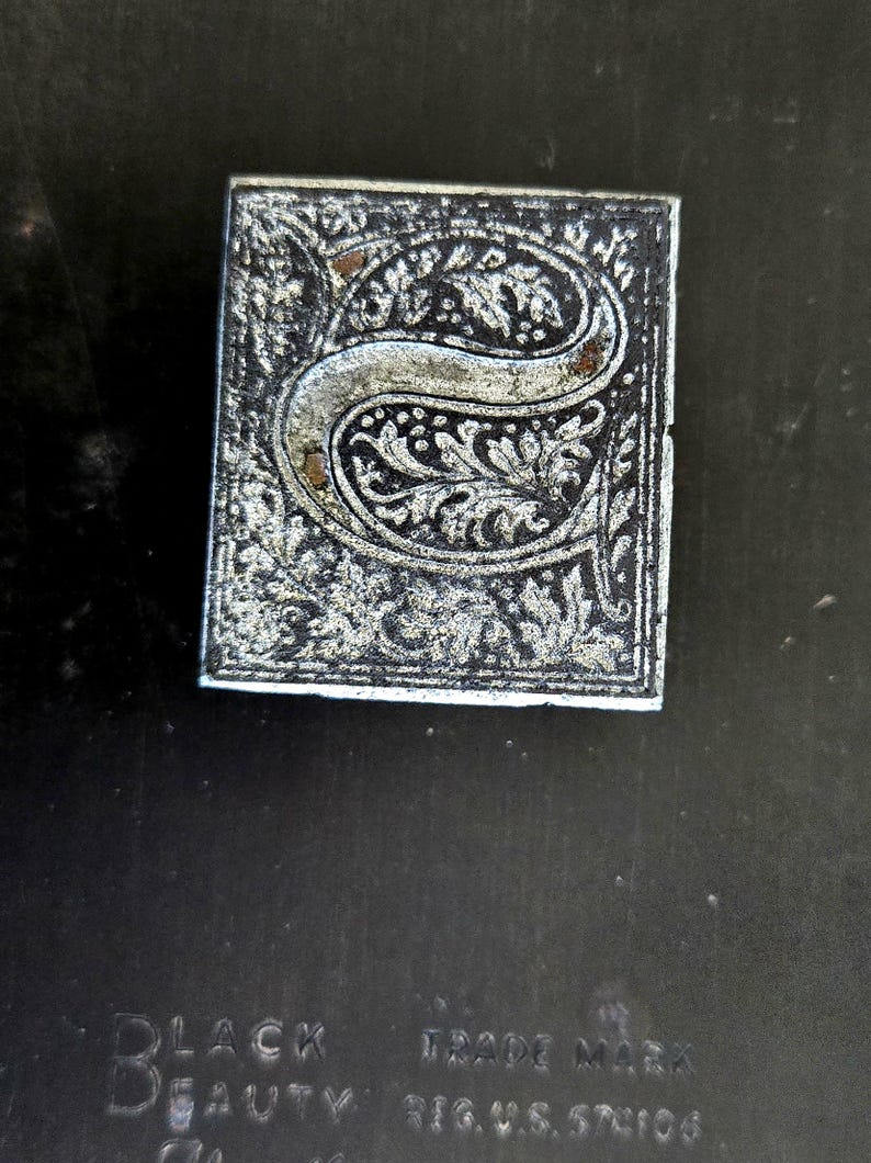 May include: A close-up of a silver metal stamp with an ornate letter "S" design. The stamp is on a black background with the text "BLACK BEAUTY TRADE MARK REG. U.S. 57106".