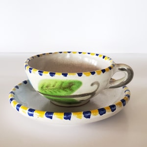 May include: A white ceramic teacup and saucer with blue and yellow trim and a green leaf design.