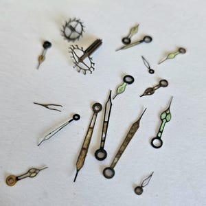 May include: A collection of vintage watch parts, including watch hands and gears, scattered on a white surface. The watch hands have a variety of shapes and colors, including gold, black, and green. The gears are metallic and intricate.