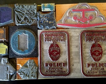 May include: A collection of vintage metal printing blocks with various designs, including a warning sign that reads "WARNING THIS AUTO IS PROTECTED BY POLICE BURGLAR ALARM SYSTEM", a Mexican food logo, and a stylized image of a bird.