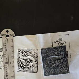 May include: A close-up of a metal stamp with a decorative letter "S" design. The stamp is on a white surface with the words "Fur Price" printed in black ink.