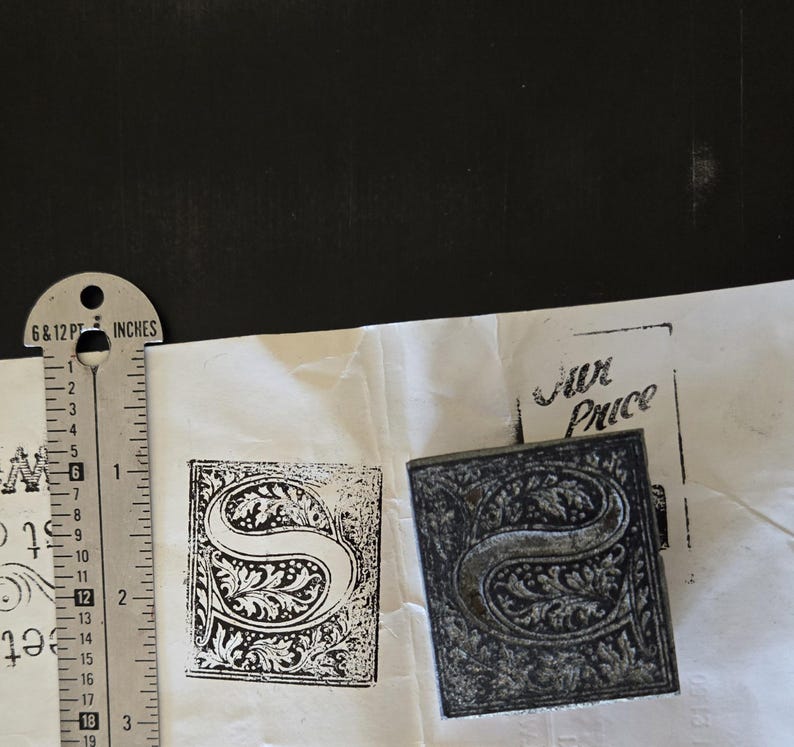May include: A close-up of a metal stamp with a decorative letter "S" design. The stamp is on a white surface with the words "Fur Price" printed in black ink.