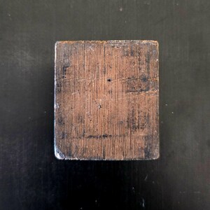 May include: A square wooden block with a dark brown finish and a wood grain pattern. The block has a slightly rough texture.