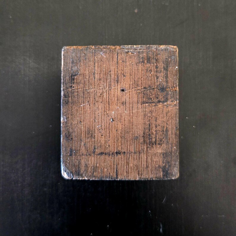 May include: A square wooden block with a dark brown finish and a wood grain pattern. The block has a slightly rough texture.