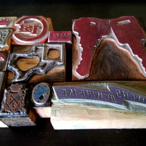 May include: A collection of vintage wood printing blocks with metal type. The blocks feature various designs, including a crown, a key, a state outline, and a company logo. The blocks are arranged in a grid pattern on a dark surface.