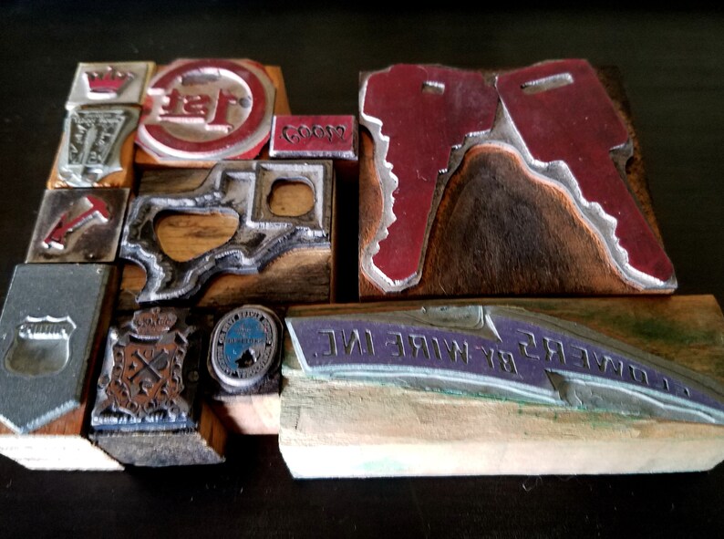 May include: A collection of vintage wood printing blocks with metal type. The blocks feature various designs, including a crown, a key, a state outline, and a company logo. The blocks are arranged in a grid pattern on a dark surface.