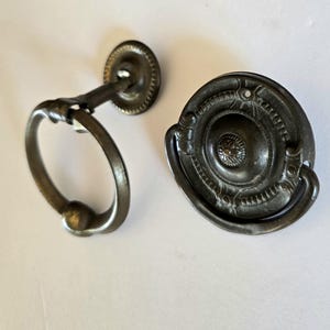 May include: Two antique bronze-toned cabinet hardware pieces. One is a ring pull with a round backplate, the other is a decorative pull with a circular design and ornate detailing. Both pieces are on a white background.