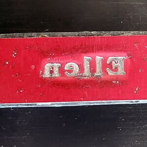 May include: A red rectangular label with a silver metallic border and the word "Ellen" printed in silver.