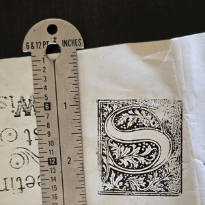 May include: A close-up of a black and white decorative letter "S" printed on white paper. The letter is surrounded by a decorative border of leaves and vines. A ruler is visible in the image, showing the letter is approximately 1 inch wide.