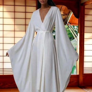 Puede incluir: Un vestido blanco fluido con mangas anchas y acampanadas y un escote en V. El vestido presenta un diseño cruzado con un cinturón a juego en la cintura. Las mangas largas crean una silueta elegante. El vestido está hecho de una tela suave y ligera.