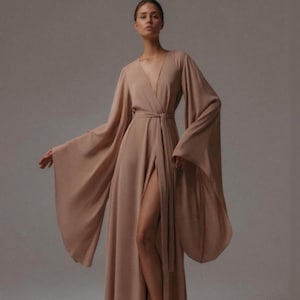 May include: A long, flowing, beige dress with a deep V-neckline and wide, bell sleeves. The dress features a high slit and a matching belt cinching the waist. The model is standing in front of a neutral background.