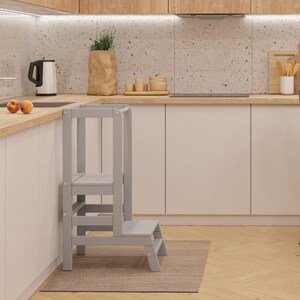 Peut inclure: Un tabouret de cuisine en bois gris clair à deux marches est placé devant un meuble blanc. La cuisine comprend des plans de travail en bois clair, un tabouret de bar gris et une esthétique moderne.