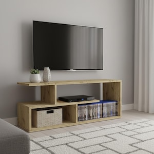 Op de afbeelding: Een lichtbruine houten tv-standaard met een modern design. De standaard heeft open planken voor opslag, waaronder een geweven mand en ruimte voor media. Een grote flatscreen-tv is erboven gemonteerd. Een grijze bank en een patroonkleed staan op de voorgrond.