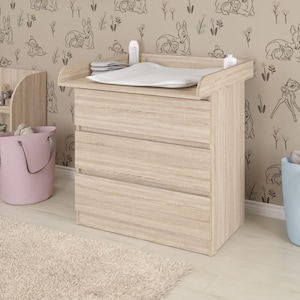 Könnte beinhalten: Eine Wickelkommode aus hellem Holz mit drei Schubladen, einer Wickelauflage und einem Regal. Eine rosa und eine blaue Aufbewahrungskiste stehen auf dem Boden. Die Tapete hat ein sich wiederholendes Muster aus Hirschen und anderen Waldtieren.