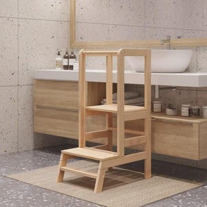 Peut inclure: Marchepied en bois clair avec une base rectangulaire, deux marches et une poignée supérieure. Le tabouret est placé sur un tapis tissé dans une salle de bain. La salle de bain comprend des meubles en bois clair et un lavabo blanc.