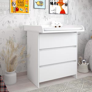 Op de afbeelding: Een witte aankleedtafel met drie lades. De aankleedtafel heeft een gebogen bovenkant en staat in een kinderkamer. De muur is versierd met ingelijste kunst en een grillig behang. De tekst "LET'S TRAVEL & ENJOY" is zichtbaar.