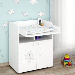 Puede incluir: Cambiador blanco con una superficie elevada y puertas de armario. Las puertas presentan un diseño de oso de dibujos animados. Una estantería encima contiene artículos para el cuidado del bebé. La pared de atrás está decorada con nubes y aviones de papel.