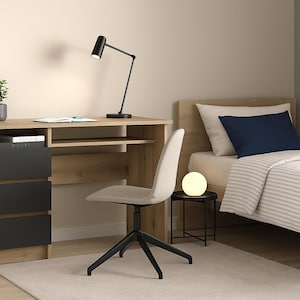 Può includere: Una scena di camera da letto moderna con una scrivania in legno chiaro con cassetti neri, una lampada da scrivania nera e una sedia bianca. Un letto con un cuscino blu navy e un tavolino con una lampada rotonda sono anche visibili.