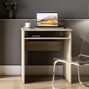 Könnte beinhalten: Ein kleiner, hellfarbiger Holzschreibtisch mit Schublade und Ablage, auf dem ein Laptop, eine weiße Kaffeetasse und ein kleines Buch stehen. Ein durchsichtiger Kunststoffstuhl und eine Stehlampe mit beigem Lampenschirm sind zu sehen. Der Hintergrund ist eine dunkelgraue Wand.