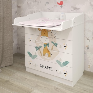 Könnte beinhalten: Weiße Wickelkommode mit Giraffen-Design. Die Kommode hat vier Schubladen mit einer Giraffen-Illustration und dem Wort "GIRAFFE". Oben liegt eine rosa Wickelauflage, und ein Mobile hängt darüber.