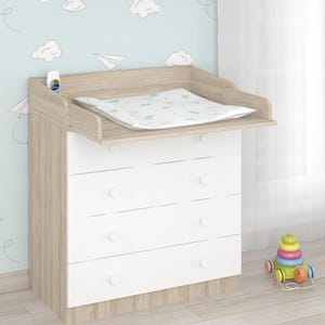 Op de afbeelding: Een licht houten en witte commode met vier lades en een aankleedkussen. De commode heeft een opstaande rand aan de bovenkant. De achtergrond is een lichtblauwe muur met wolken- en papieren vliegtuigstickers.