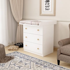 Könnte beinhalten: Weiße Wickelkommode mit drei Schubladen und goldfarbenen Knöpfen. Die Oberseite hat eine Wickelauflage mit Muster. Ein gerahmter Druck mit dem Text "cool kids never sleep" hängt an der Wand.