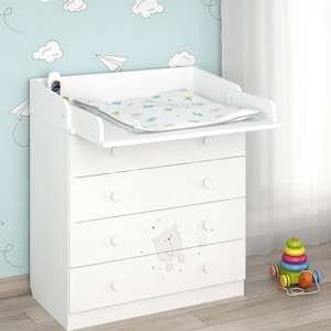 Op de afbeelding: Witte commode met vier lades en een aankleedkussen. De lades hebben ronde knoppen en een berenafbeelding. Een kleurrijk speelgoed staat op de vloer naast de commode. De muur erachter is lichtblauw met wolken- en papieren vliegtuigdecals.