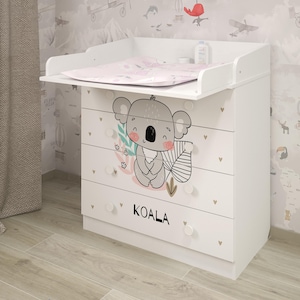 Può includere: Cassettiera bianca con grafica di koala e la scritta "KOALA" sui cassetti. Il fasciatoio include un materassino e una giostrina con giocattoli colorati appesi. I cassetti hanno pomelli rotondi bianchi.