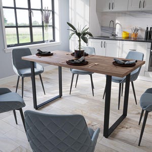 Peut inclure: Une salle à manger moderne avec une table rectangulaire en bois et des pieds en métal noir, entourée de six chaises grises rembourrées. La table est dressée avec des assiettes noires et des couverts. La pièce a de grandes fenêtres.