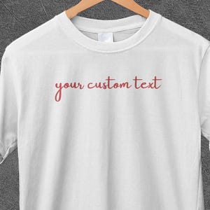 Può includere: T-shirt bianca con la scritta "your custom text" in corsivo rosso. La maglietta è appesa a una gruccia di legno su sfondo grigio. La maglietta è a girocollo.