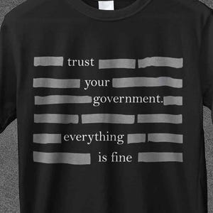 Può includere: T-shirt nera con la scritta "trust your government, everything is fine" in lettere grigie. La maglietta è appesa a una gruccia di legno su uno sfondo grigio scuro. Il marchio "JoJoTeez" è in alto.