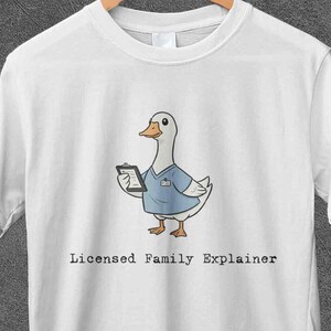 May include: White t-shirt featuring a cartoon goose wearing a blue medical uniform and holding a tablet. The text "Licensed Family Explainer" is printed below the goose. The shirt is hanging on a wooden hanger.