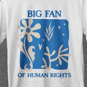 Peut inclure: T-shirt blanc avec un motif bleu qui dit "BIG FAN OF HUMAN RIGHTS". Le motif représente une fleur, des feuilles et d'autres formes. Le t-shirt est suspendu à un cintre en bois.