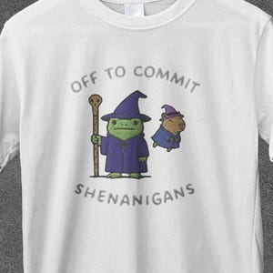 Może przedstawiać: Biała koszulka z napisem "Off to Commit Shenanigans" i kreskówkowym czarodziejem oraz małą istotą. Czarodziej jest zielony, nosi fioletową szatę i kapelusz, trzymając laskę. Druga istota jest niebieska.