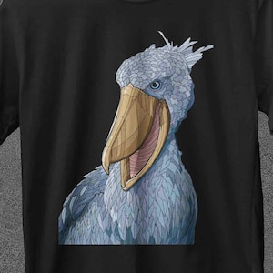 May include: A black t-shirt with a Shoebill stork illustration. The bird is depicted with blue and grey plumage and a prominent beak. The brand name "JoJoTeez" is visible at the top left. The shirt is hanging on a wooden hanger.